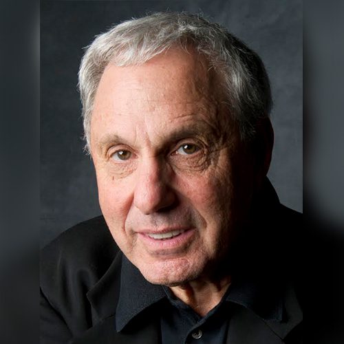 Older man with gray hair wearing a black shirt and jacket, looking directly at the camera against a dark background.
