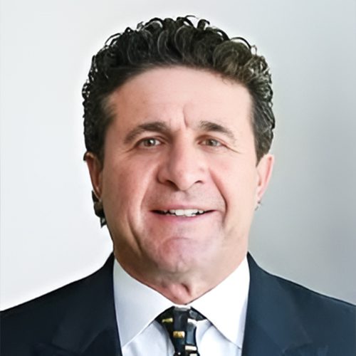 Paul Schiada Photo-Picsart-AiImage Smiling man in a suit with curly hair, wearing a patterned tie, against a plain background.