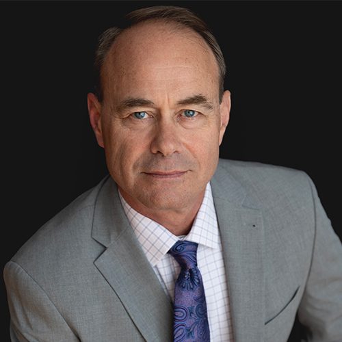 Robert Mann head shot 2024 Man in a grey suit and blue tie against a dark background, looking directly at the camera.